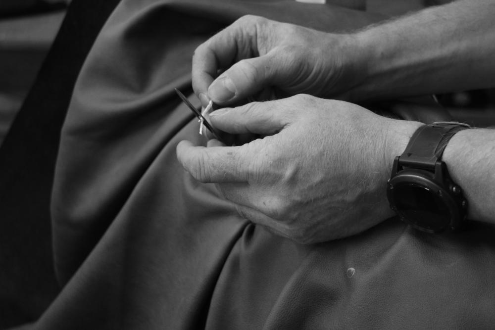 Black and white close-up of an upholsterer's hands threading twine through a long upholstery needle over dark leather in The English Sofa Company's Manchester workshop.
